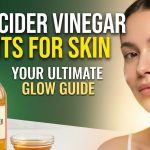 A bottle of raw apple cider vinegar sitting next to fresh red apples and cotton skincare pads on a wooden table.